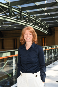 An image of Rachel Parry standing on the second floor of a building, next to glass balustrade. Sun is shining through a glass roof.
