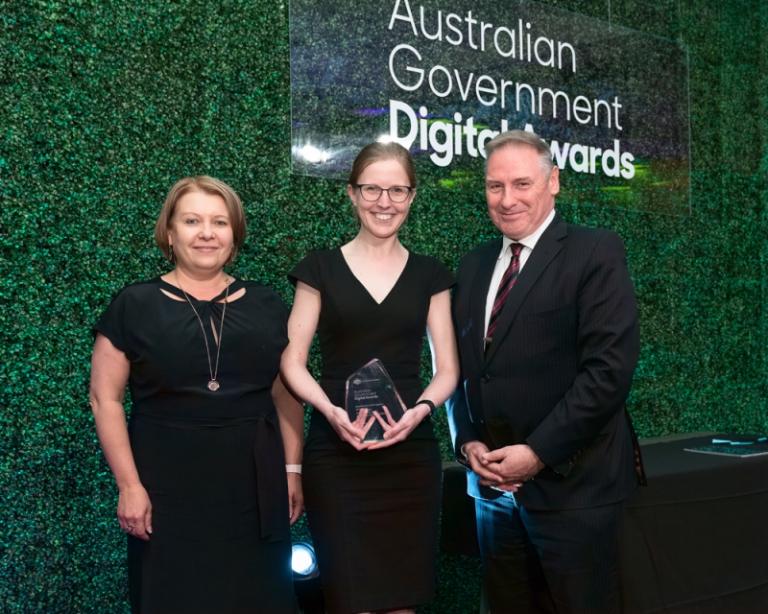 Two women and a man in formal attire stand in front of a sign that reads ‘Australian Government Digital Awards’. The women in the centre is holding a clear glass trophy.
