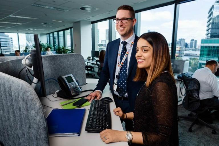 A man and a woman in office attire stand at a workstation in an office.