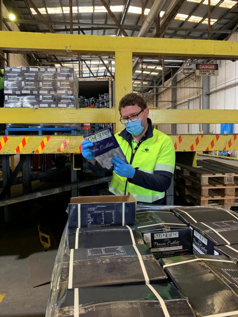 A man wearing high-vis clothing, a face mask and gloves, inspecting a packet of prawn cutlets from a pallet in a warehouse