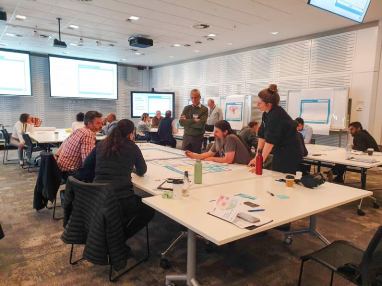  Men and women are standing and sitting around tables, discussing work on documents and screens.