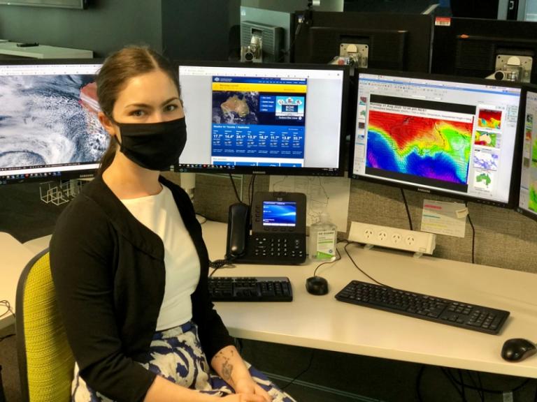 A women wearing a mask, sitting at a desk in front of weather images on screens