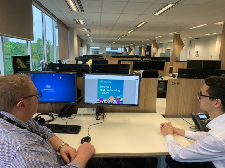 Two men dressed in business attire sit (socially distanced) apart at a desk, viewing the cover page of the Driving a High-performing Culture Framework on a computer screen.