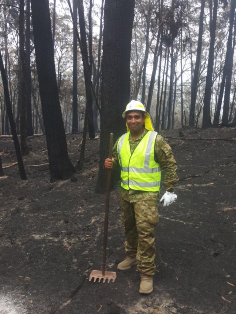 A man wearing a high-vis vest and hard hat over an Australian Army camouflage uniform, standing amidst fire blackened trees and ground, holding a metal rake.
