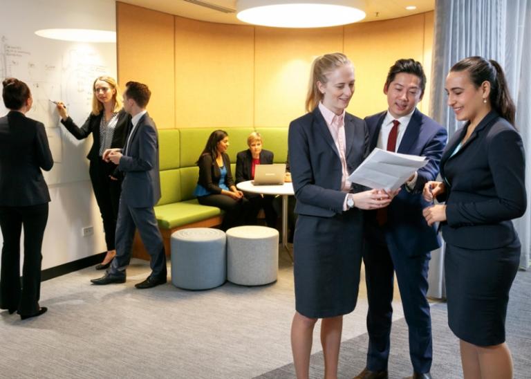 Men and women in office attire standing and sitting in a workplace, discussing work on a whiteboard, document, and laptop