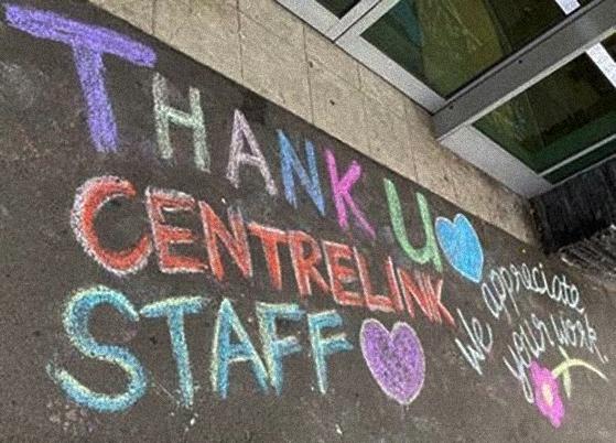 Colourful chalk drawing with writing that reads ‘thank u Centrelink staff, we appreciate your work’ with love hearts and a flower, on a footpath in front of glass sliding doors.