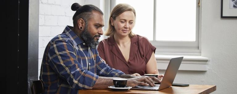 A stock photo of two people in an office environment looking at the same laptop screen. A male-presenting human has a man bun and a plaid shirt, while a female-presenting human wears a maroon top.