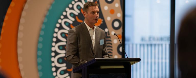 Image of Gary Rake standing at a lectern presenting to a crowd. Behind him is Aboriginal artwork.