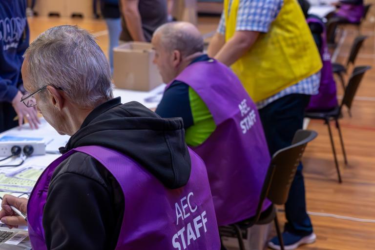 A photograph of AEC polling booth staff registering voters at a desk on election day. 