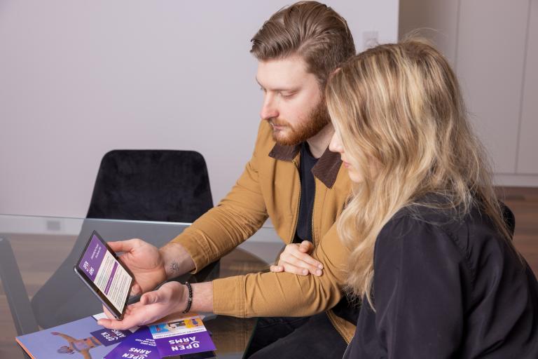 A photograph of a couple sitting at a table, looking at an online mental health platform on an iPad, with serious expressions on their faces.  