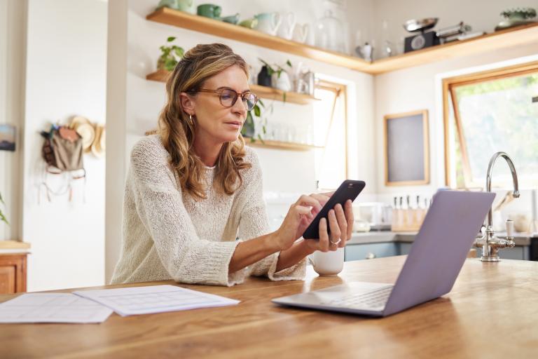A photograph of a woman sitting at her kitchen bench, completing her tax return on her phone. 