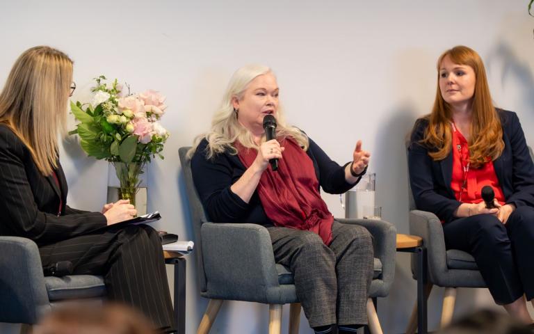 Three people sitting in chairs, the person in the middle is holding a microphone and speaking to a crowd (not pictured)