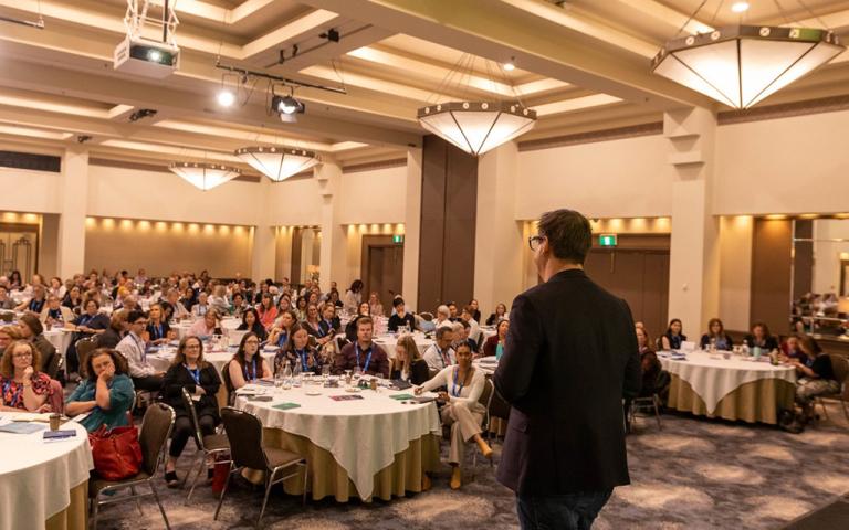 A person standing in front of a crowd who are sitting at round tables