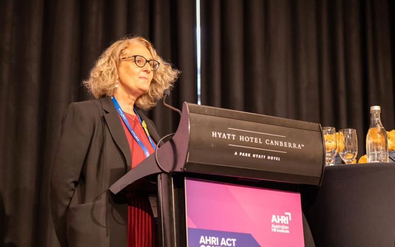 A person, Paula Goodwin, standing at a lectern on a stage, talking to an audience through a microphone. The audience is not pictured.