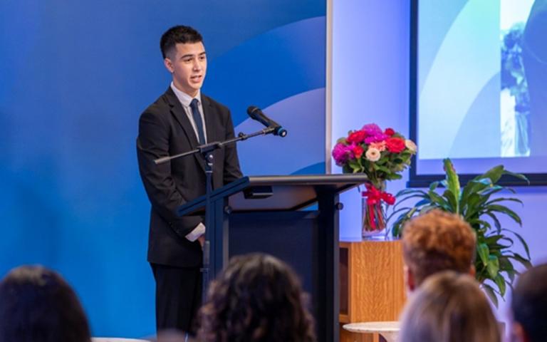 a person standing at a lectern on stage in front of a crowd, speaking into a microphone