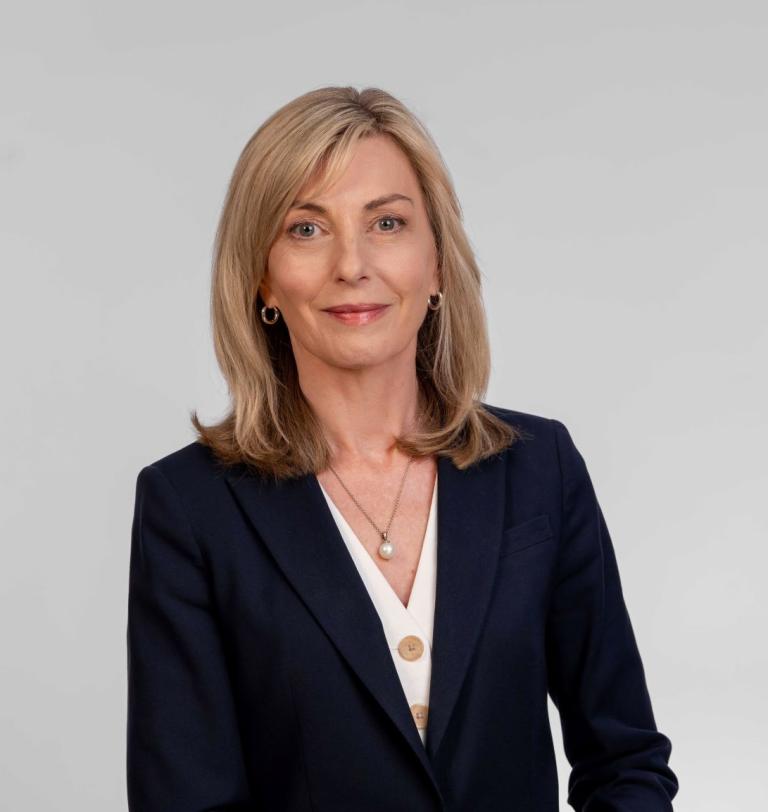 This is a headshot of Australian Public Service Commissioner Jacqui Curtis PSM. She stands in front of a flat grey background wearing a dark coloured jacket and white shirt with blonde hair down to her shoulders