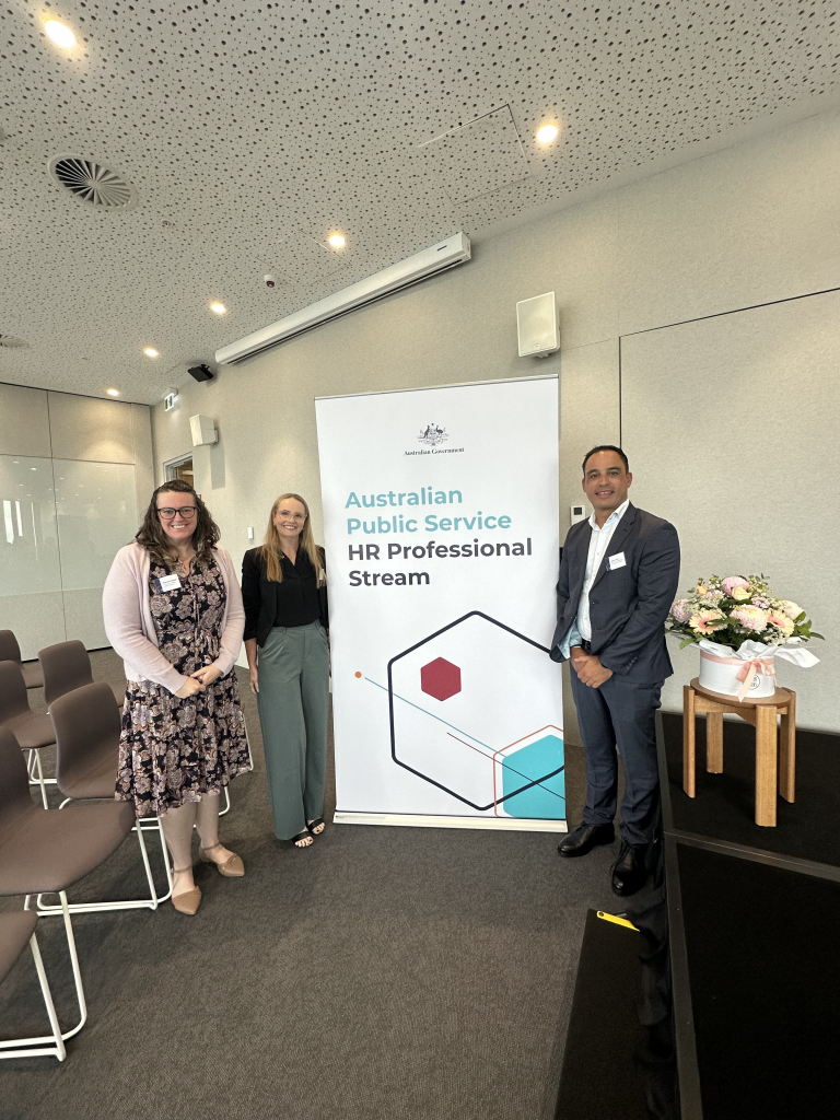 Three HR professionals in Brisbane smile in from of a HR Professional Stream banner