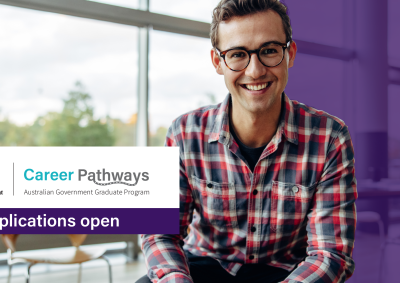 A young man wearing glasses and a checked shirt sits in an office smiling at the camera. Text reads: “Australian Government Career Pathways. Applications open.”
