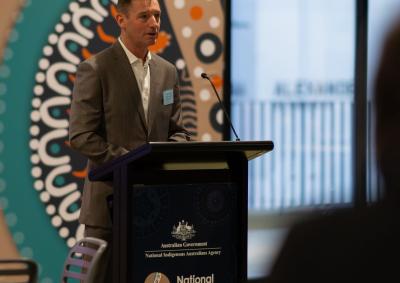 Image of Gary Rake standing at a lectern presenting to a crowd. Behind him is Aboriginal artwork.