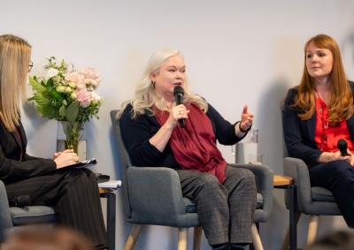 Three people sitting in chairs, the person in the middle is holding a microphone and speaking to a crowd (not pictured)