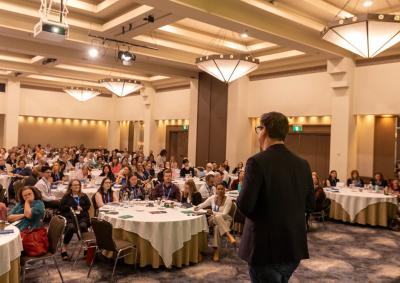 A person standing in front of a crowd who are sitting at round tables
