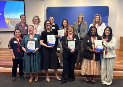 Alternative text: a group of 12 people standing on and in front of a small platform, holding certificates and smiling at the photographer.