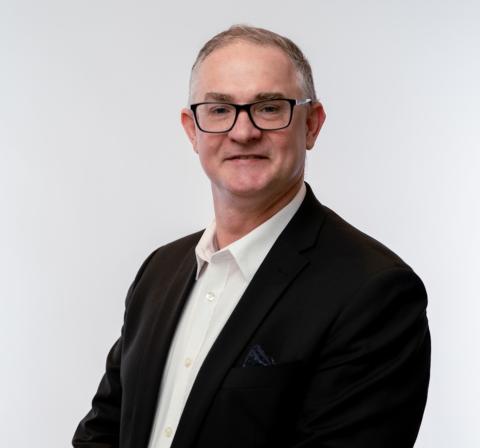 A head shot photograph of Damien Booth. A man smiling wearing glasses and a black blazer.