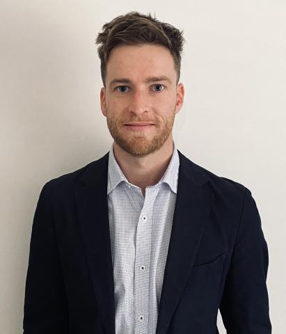 Photograph of Charlie Middleton APSC Assistant Commissioner with short brown hair, wearing a white checkered shirt and a dark blazer, standing against a plain light-colored background. 