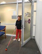 Woman walking through a door wearing black jumper, red pants, and white shoes, using a walking stick.