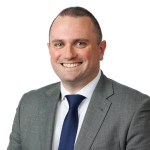 a photo of Tim Abrahams showing him from his chest upwards. He is wearing a grey suit with a navy blue tie and white shirt. He is smiling and looking directly at the camera.