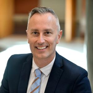 a photo of Scott Mischke showing him from his chest upwards. He is wearing a black suit with a light-blue and purple tie and white shirt. He is smiling and looking directly at the photographer.