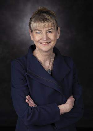 portrait of Justine Greig, a woman smiling, posing with folded arms against a dark background. 